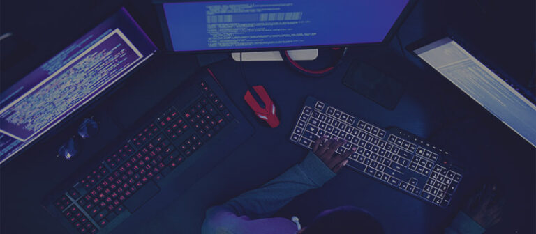Una persona que trabaja en una computadora con pantallas duales en una habitación oscura, lo que sugiere un entorno que podría estar asociado con programación o actividades cibernéticas.