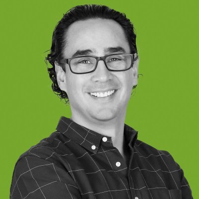Retrato de un hombre sonriente con pelo rizado hasta los hombros, gafas y camisa a cuadros, sobre un fondo verde brillante con Hans Hatch.