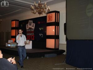 Un hombre habla por un micrófono en un podio, con un fondo de escenario iluminado con pancartas del evento y una pantalla grande a la derecha, destacando las estrategias de marketing de Hans Hatch.