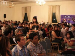 El público en una sala de conferencias escucha atentamente al orador Hans Hatch, algunos se giran para mirar a la cámara, con filas de sillas y pancartas del evento al fondo.