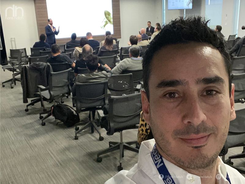 Hombre tomándose un selfie en una sala de conferencias con una audiencia escuchando a un orador de fondo.