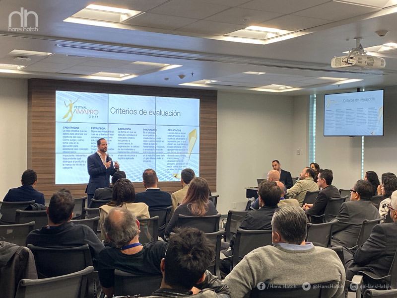 Hombre haciendo una presentación ante una audiencia en una sala de conferencias con una pantalla de proyección que muestra diapositivas sobre los criterios de evaluación.
